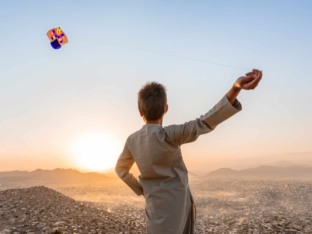 A boy flying a kite on a hilltop above Kabul at sunset